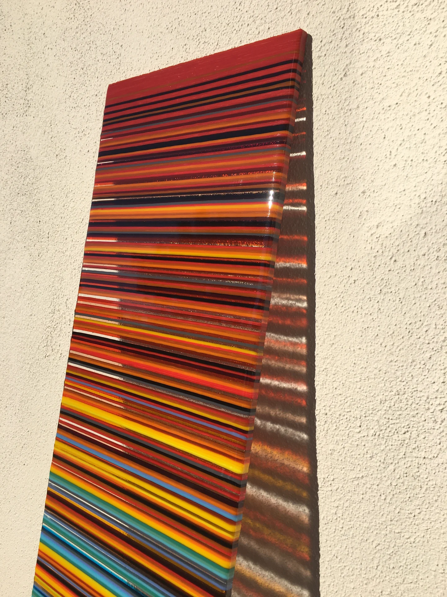 A tall narrow striped panel in red, orange, yellow and blue stands against a pale textured wall.