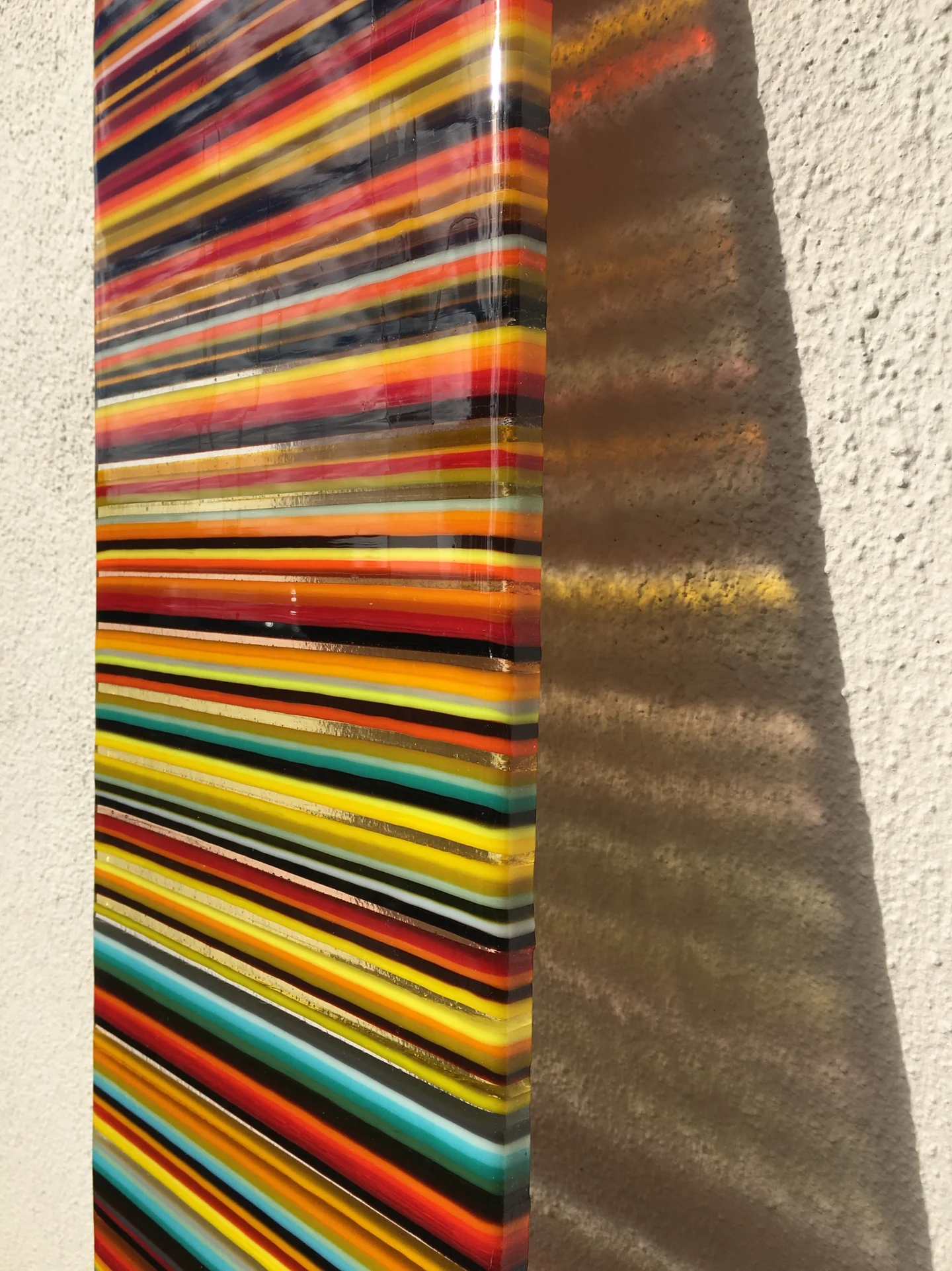 Close angled view of narrow red, orange, yellow and blue stripes beside a dark vertical edge and striped shadow on a pale wall.