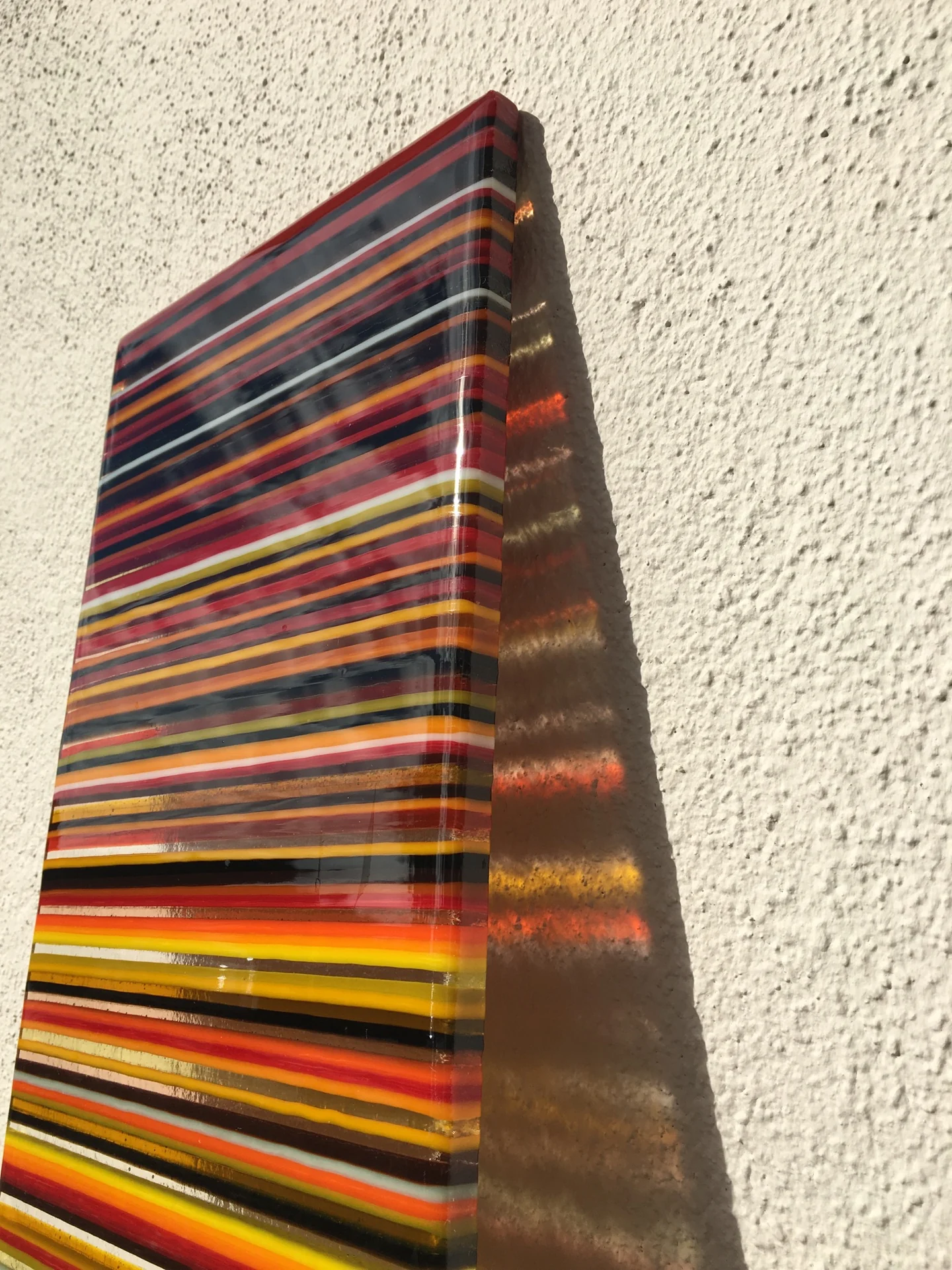A low angled close-up shows red, orange, yellow and dark horizontal stripes on a tall panel beside a pale textured wall.