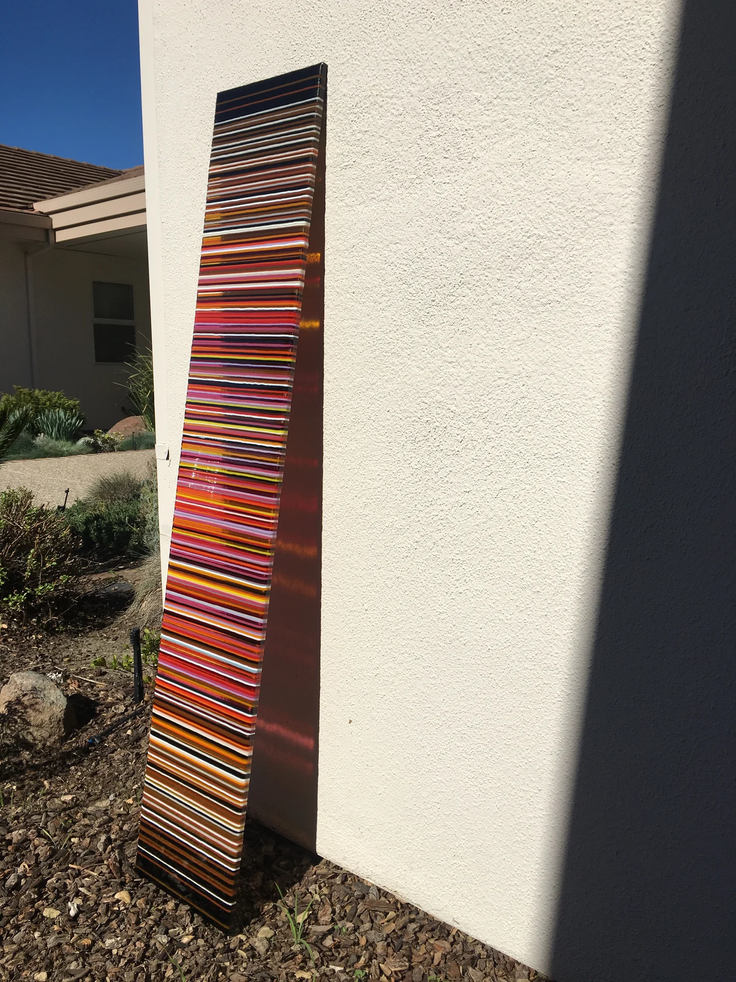 A tall panel with narrow red, orange and dark horizontal stripes stands beside a pale exterior wall.