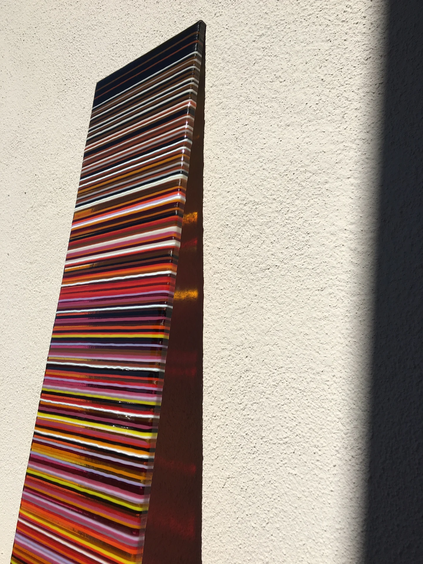 An angled view of a tall, warm-coloured striped panel beside a pale wall and a broad dark shadow.