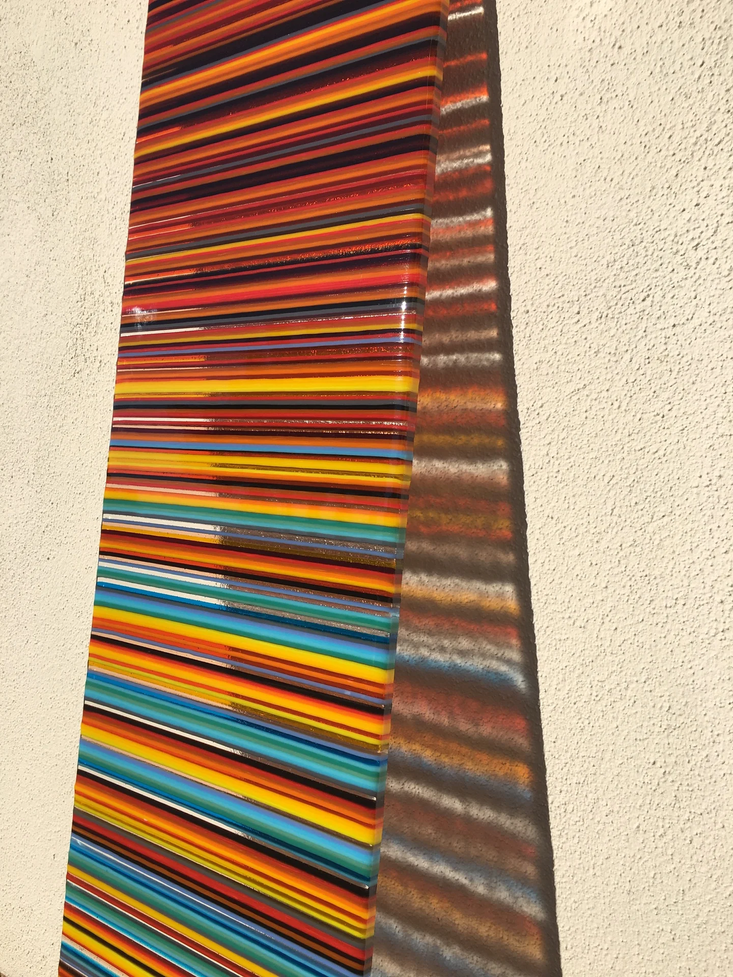 A tall narrow panel of red, orange, yellow, blue and turquoise horizontal stripes stands against a pale textured wall.