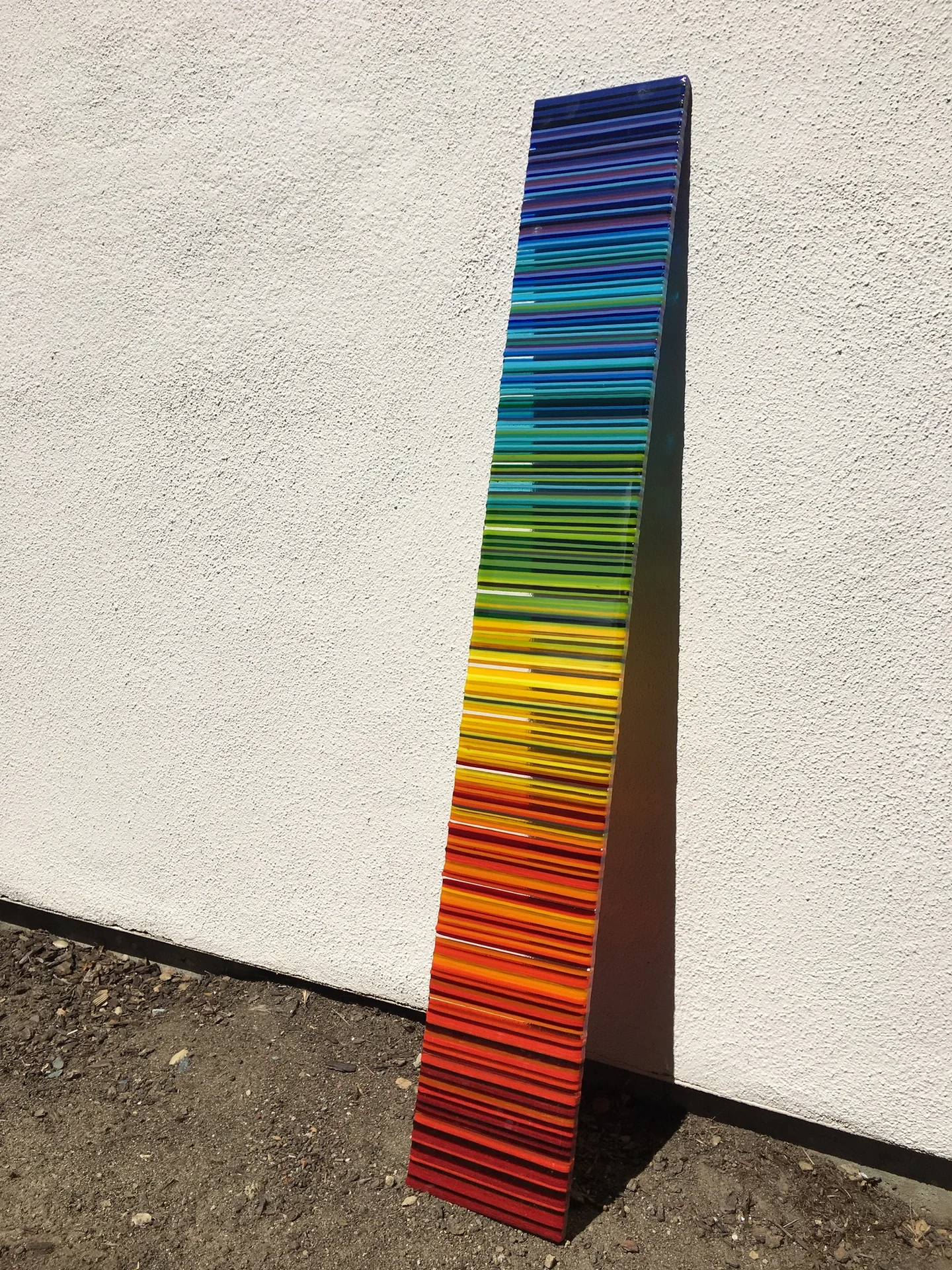 A narrow upright panel stands against a pale wall, its horizontal stripes shifting from blue and green at the top through yellow and orange to red below.