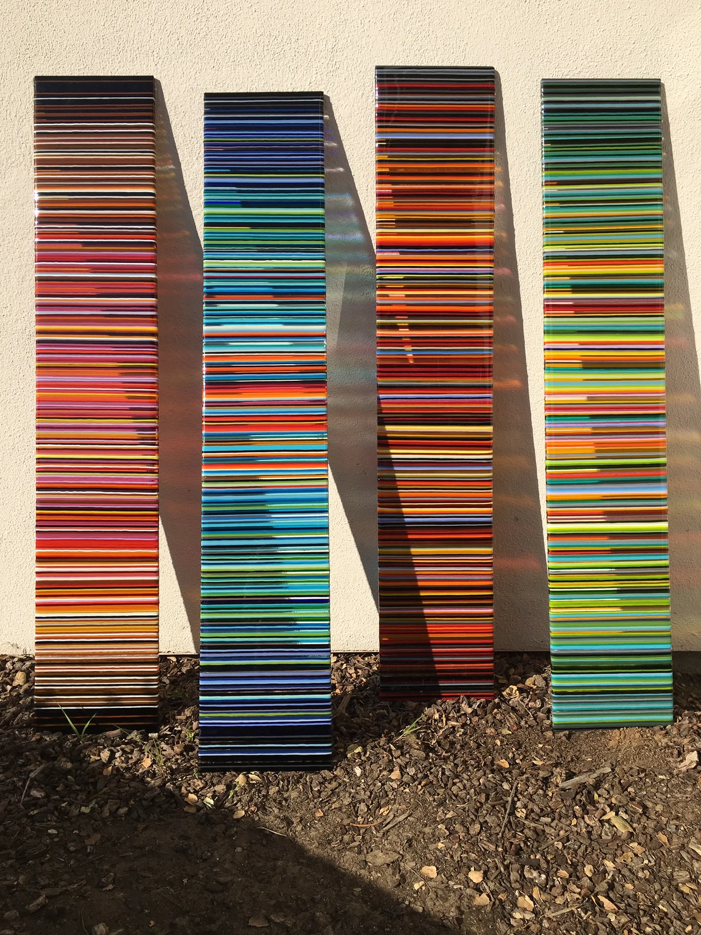 Four tall narrow panels stand in a row against a pale wall, with alternating warm red-orange and cool blue-green stripes.