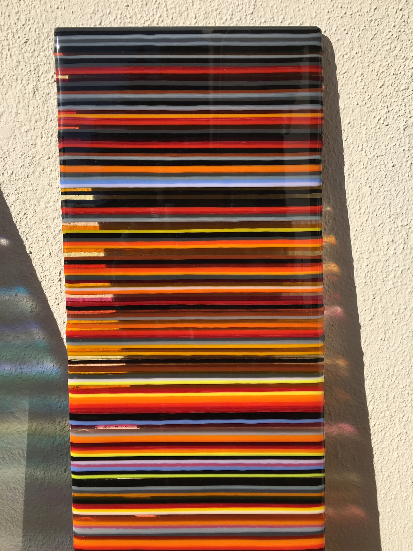 A front-facing tall panel has narrow dark, red and orange horizontal stripes with yellow, blue and white bands between them.
