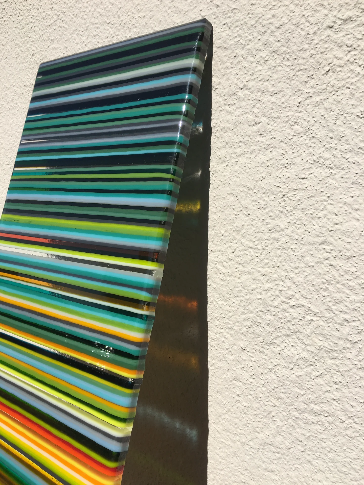 A close angled view of a narrow panel with blue, turquoise, green and yellow horizontal stripes against a pale wall.