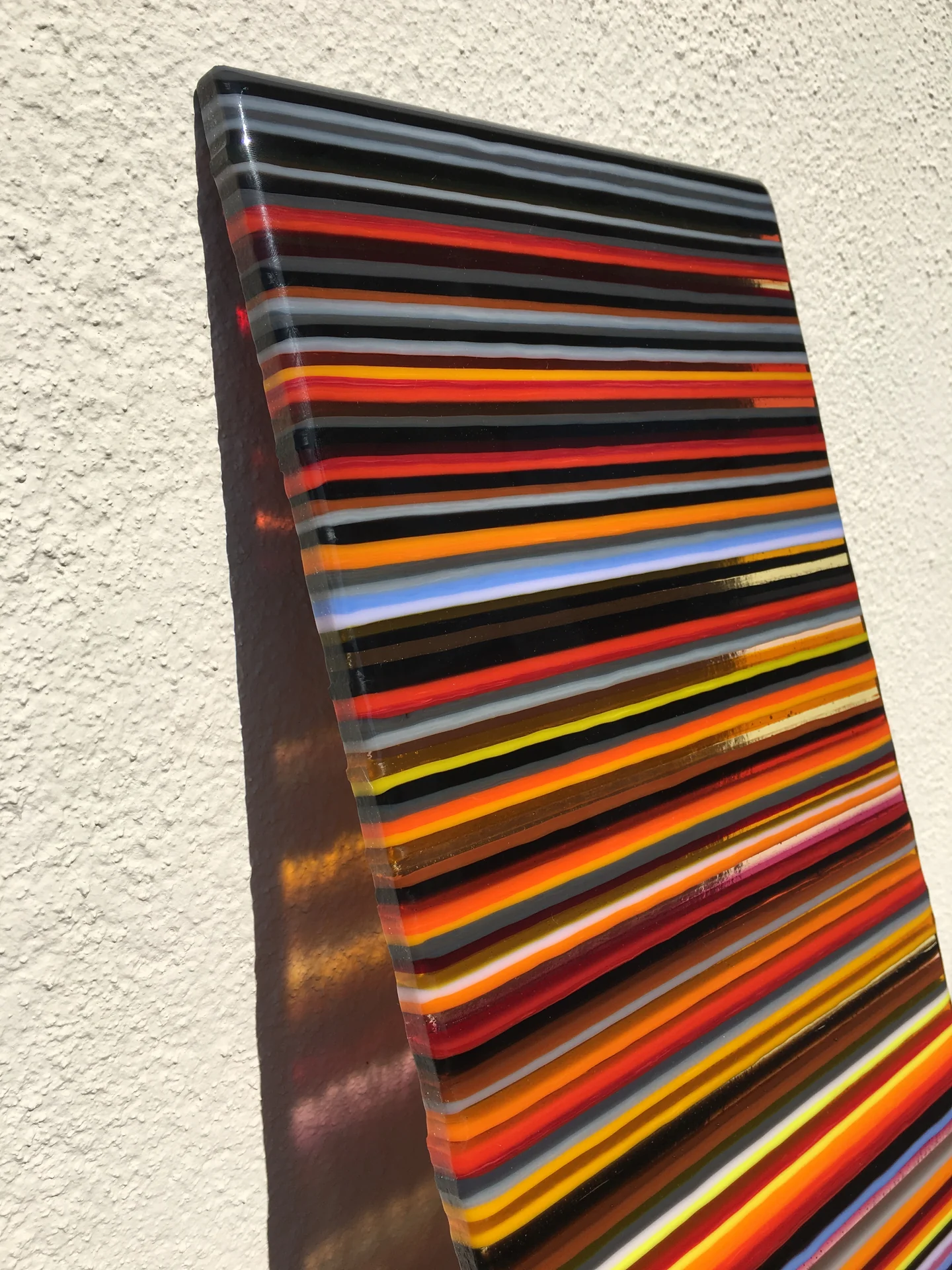 A low angled view of a tall panel with dark, red, orange and yellow horizontal bands against a pale wall.