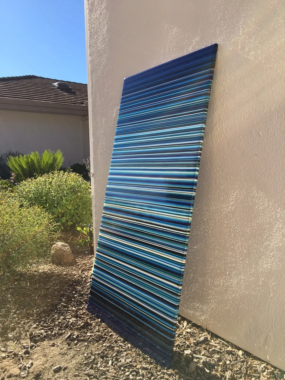 A tall blue-striped panel leans against a pale exterior wall, with shrubs and gravel visible beside it.