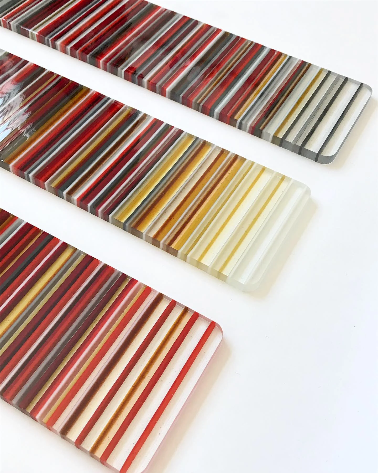 Three long rectangular striped forms lie diagonally across a white background, shifting from dark reds and grays to yellow and pale cream.
