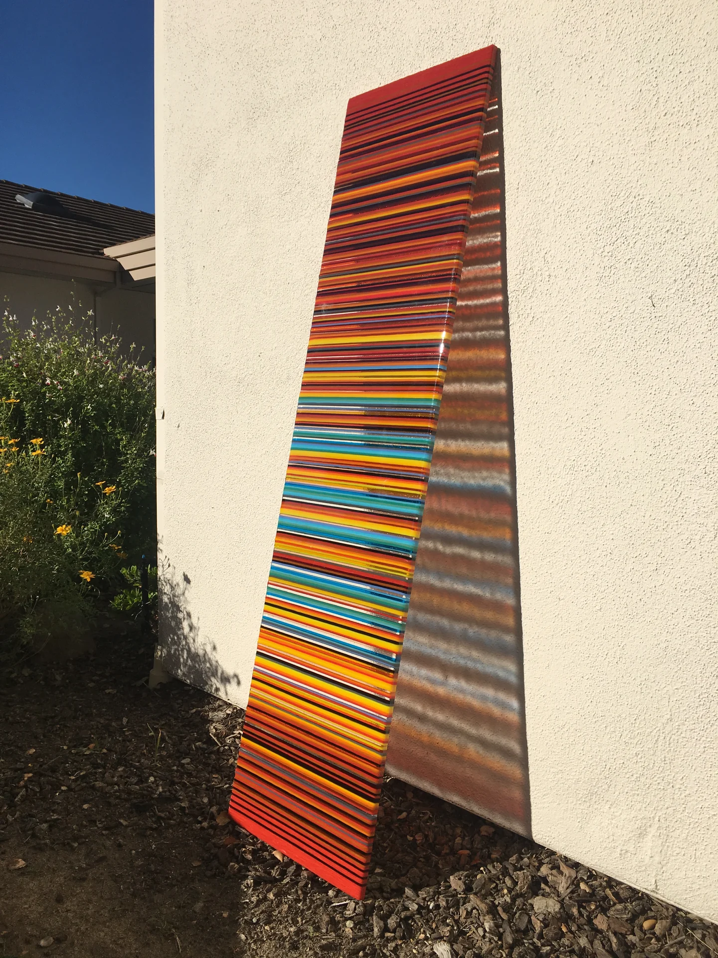 A tall narrow panel with red, orange, yellow and turquoise stripes stands against a pale exterior wall above gravel.