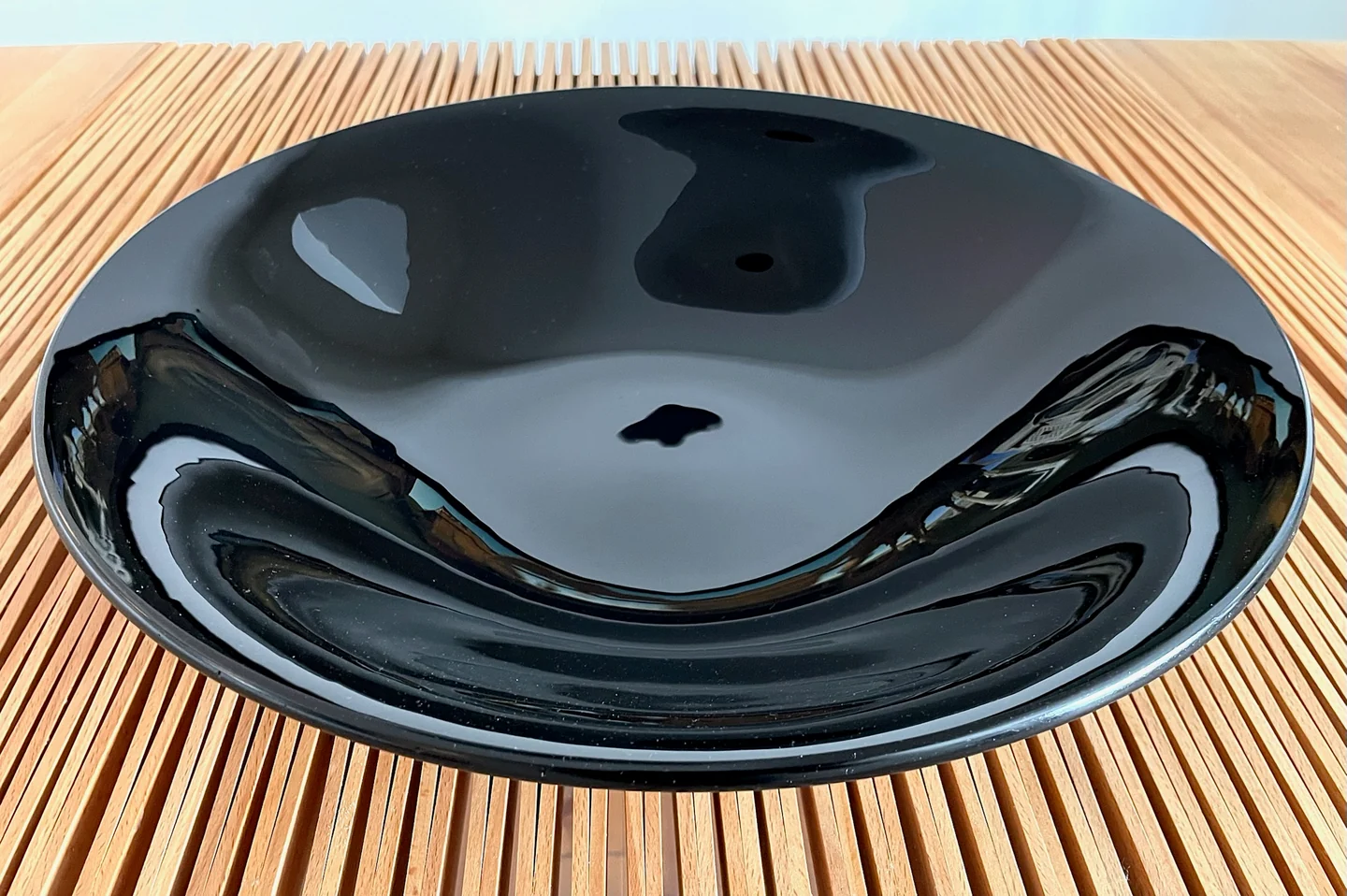 A glossy black bowl with a curved reflective interior sits on a slatted surface, seen at an angle.