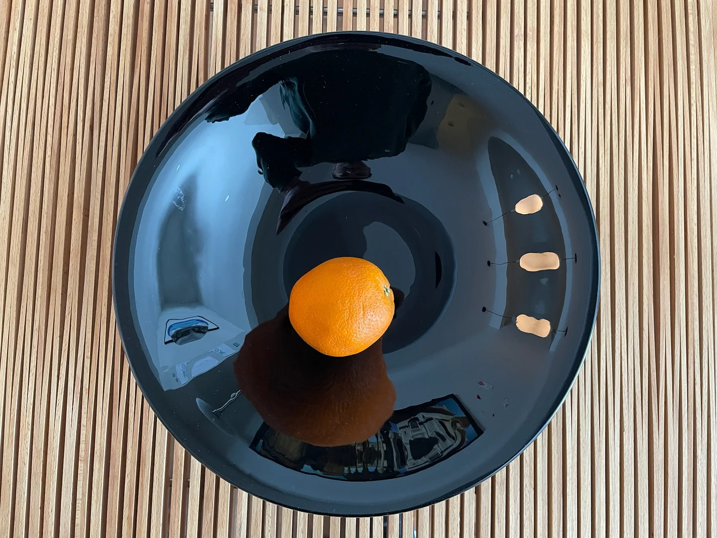 Seen from above, a glossy black bowl holds an orange near its center on a closely slatted surface.