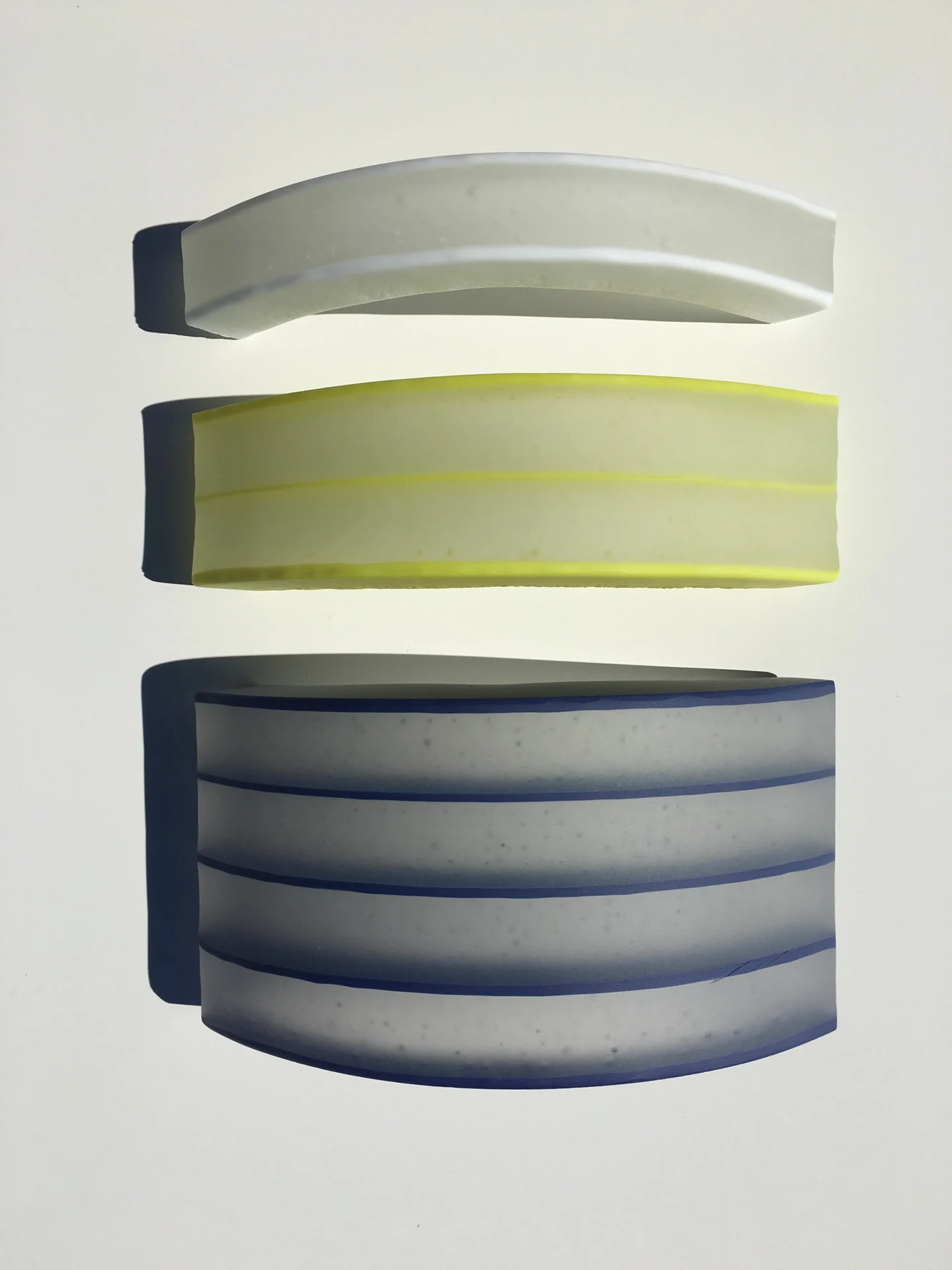 Three separate curved forms in pale gray, yellow-green and blue-gray are arranged vertically on white.