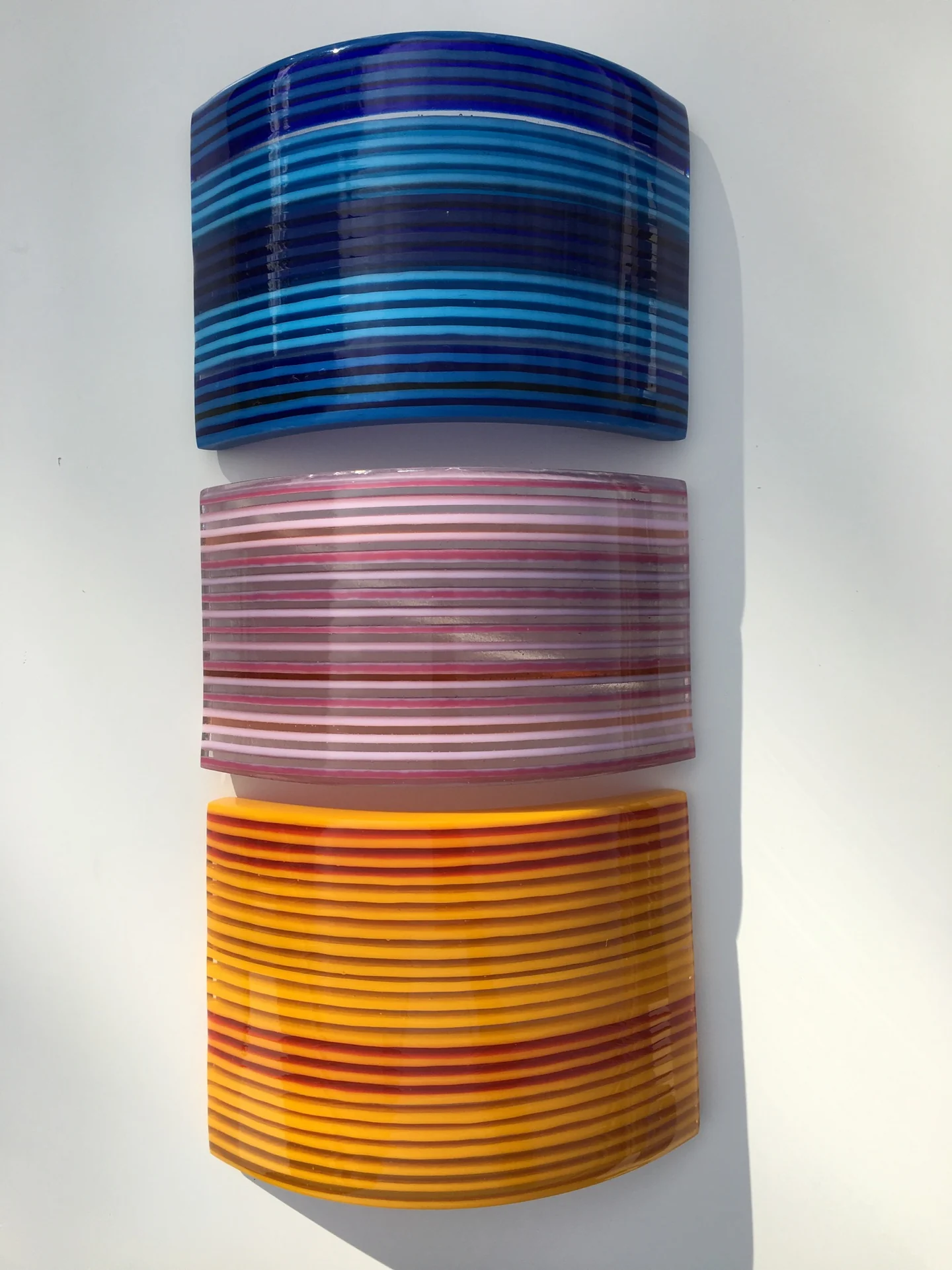 Three curved horizontally striped forms in blue, mauve and golden yellow are stacked one above another on white.