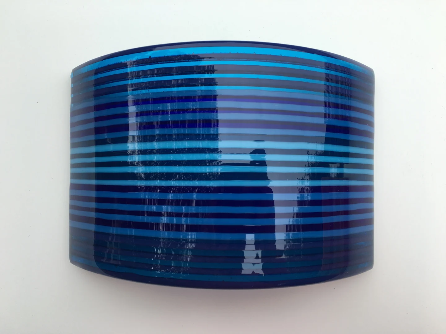 A single broad curved form has dense horizontal bands in dark and bright blues against a pale background.