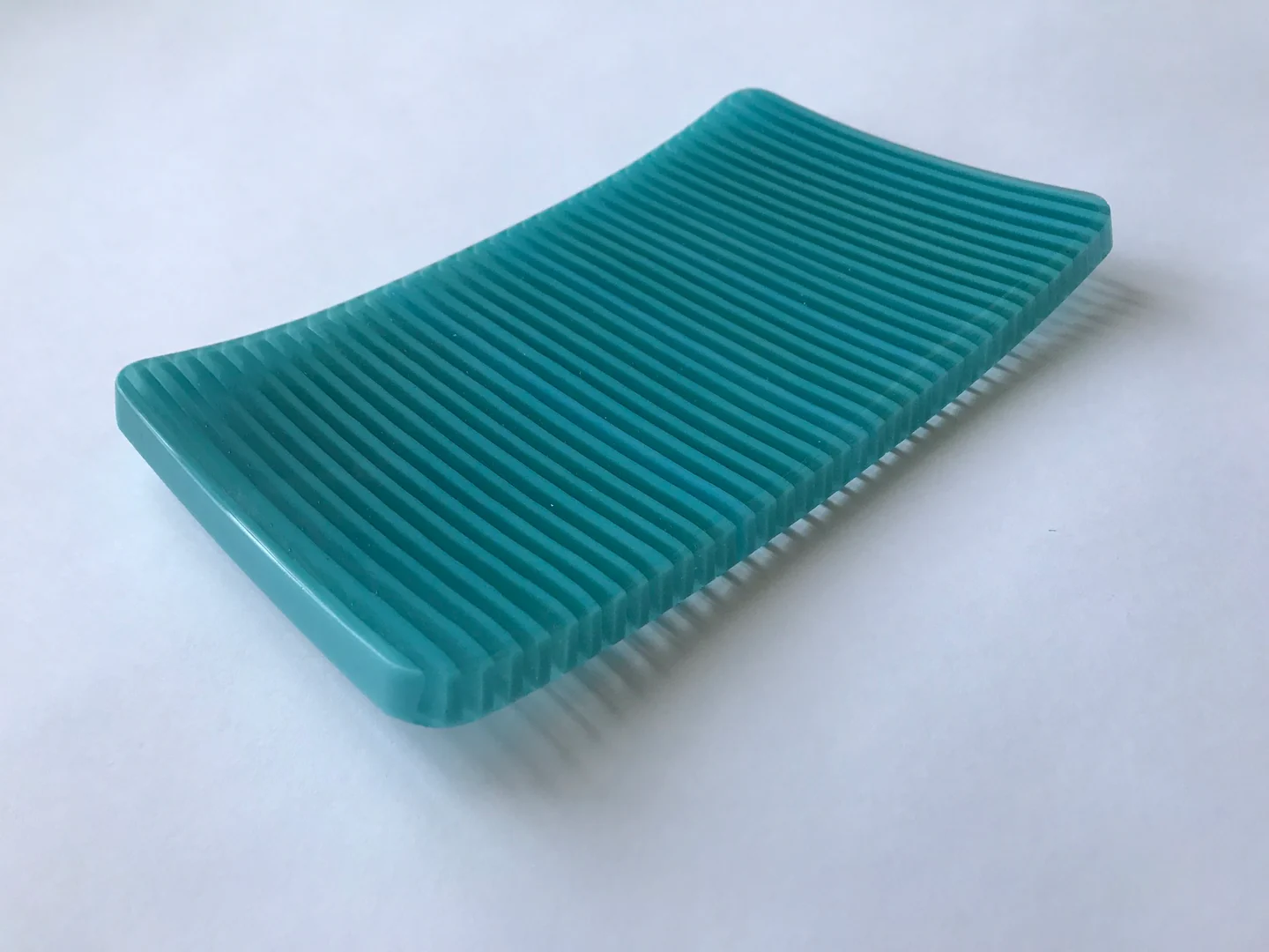 A shallow turquoise curved form has fine parallel ridges and is photographed diagonally against a pale background.