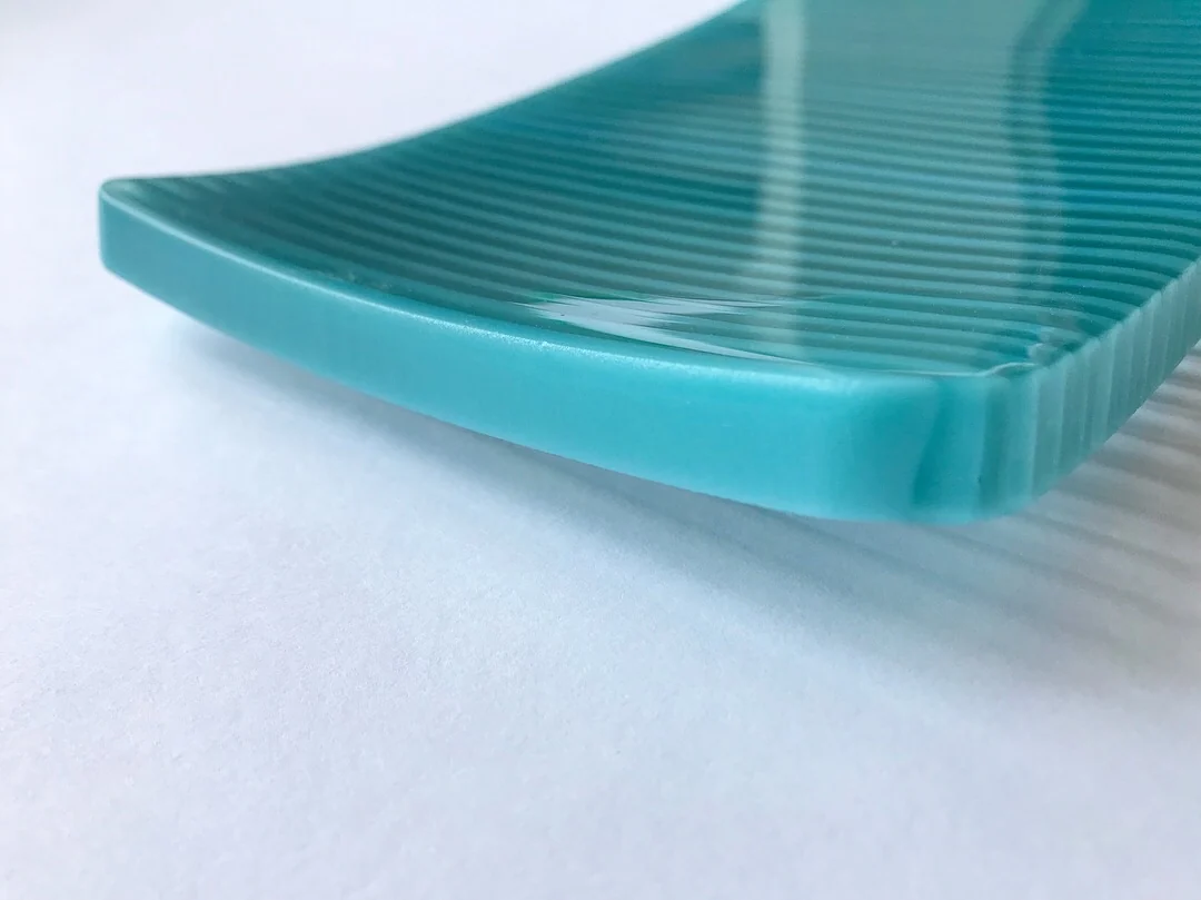 A close side view of a turquoise curved form shows fine ridges running toward its raised edge on white.