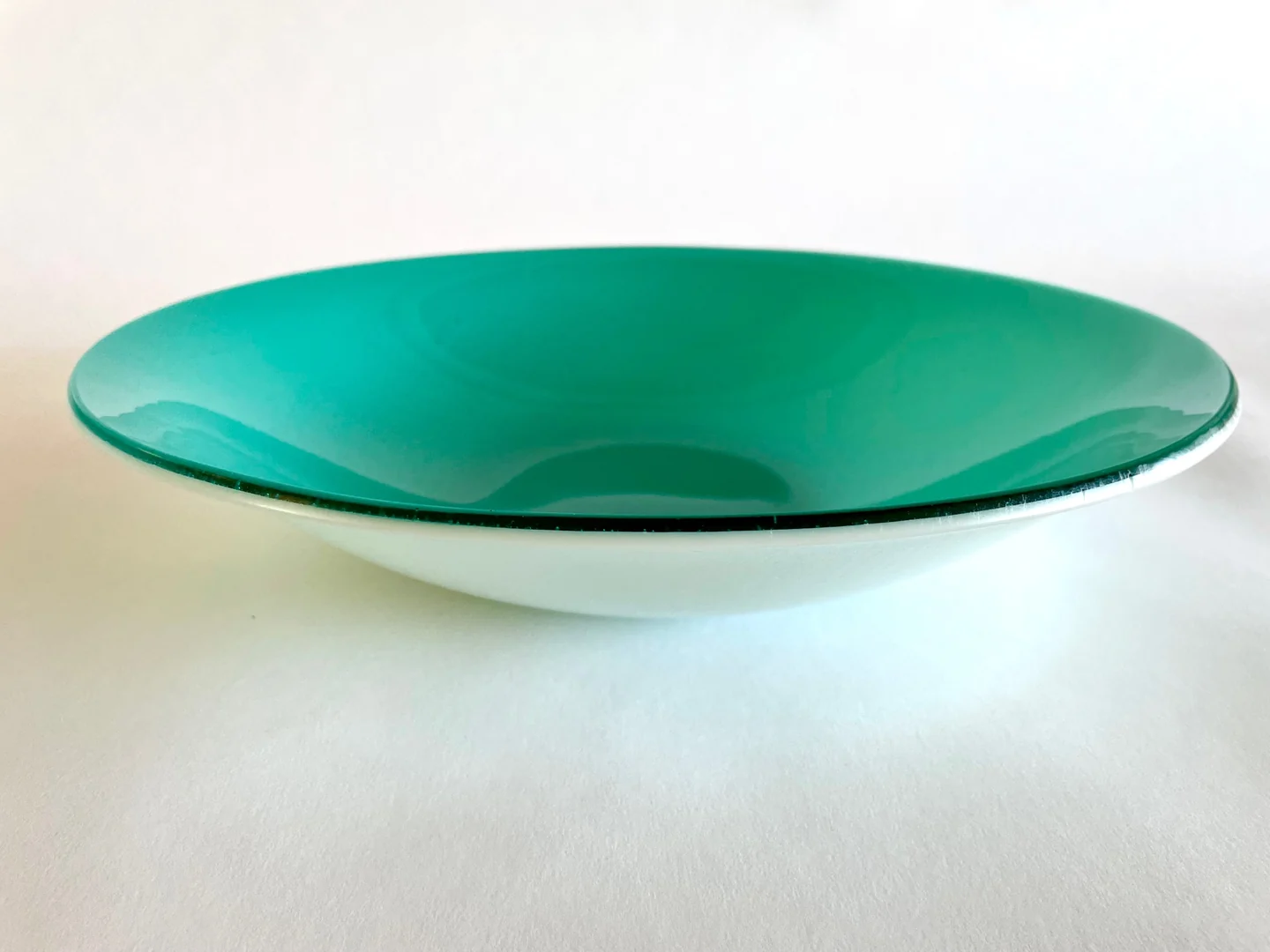 A shallow round aqua form with a darker central depression is seen at a low angle on a white surface.