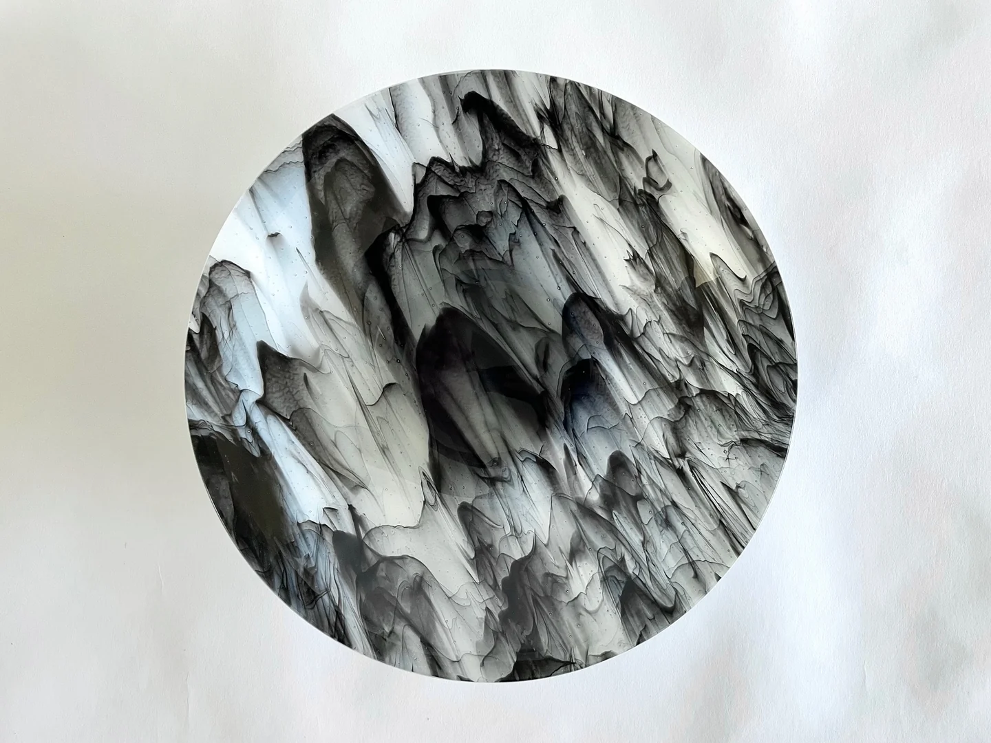 A circular form seen from above is filled with swirling black, gray and white marks on a pale background.
