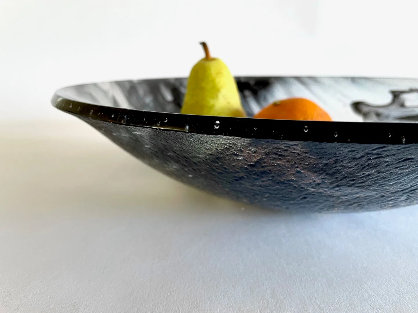 A low side view shows a wide black-and-white marbled shallow form holding a green pear and an orange.