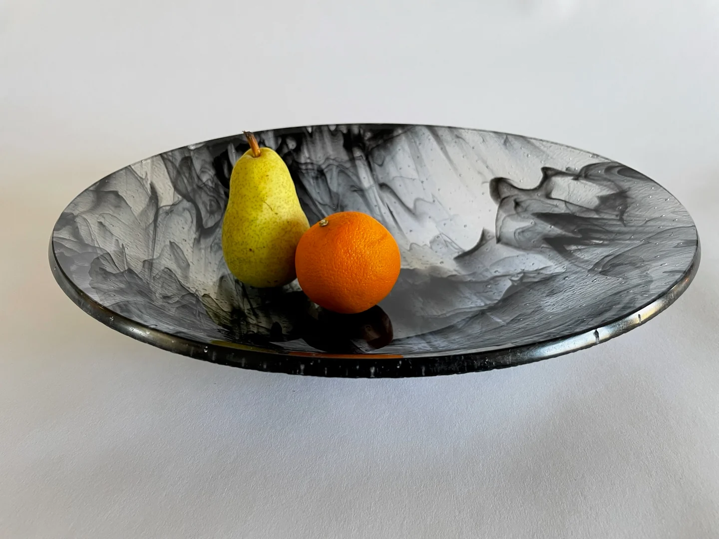 A wide black-and-white marbled shallow form holds a green pear and an orange against a pale background.