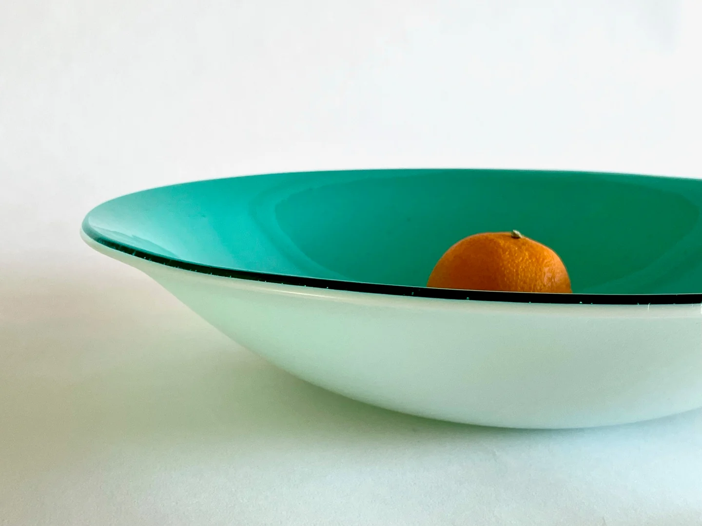 An aqua-lined shallow form with a white exterior holds a single orange against a pale background.