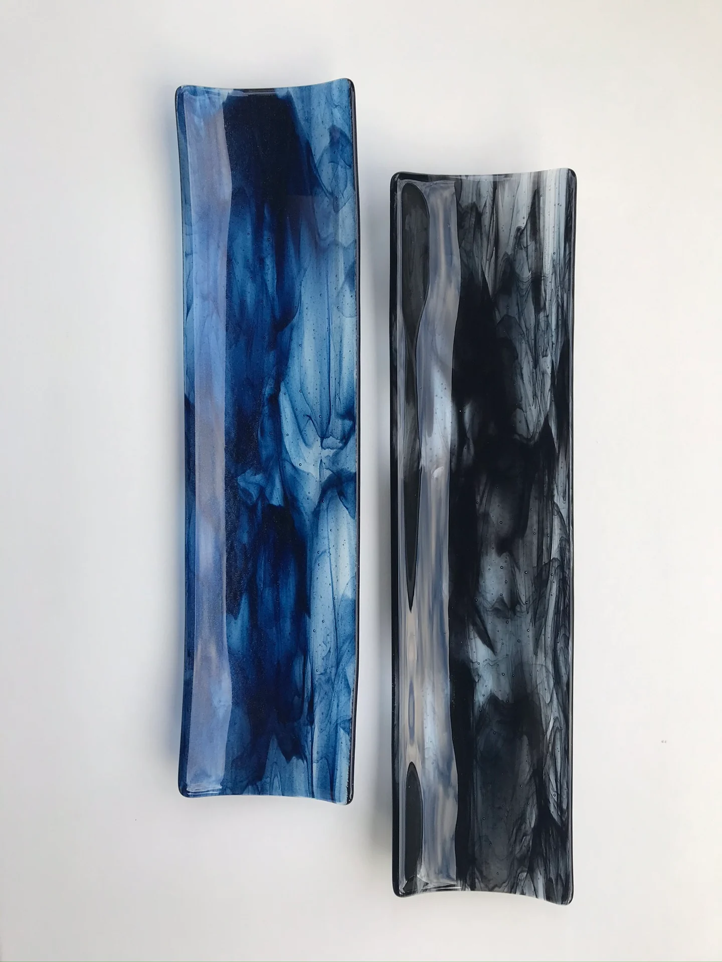Two narrow upright forms with curved ends stand on white, one streaked blue and the other gray and black.