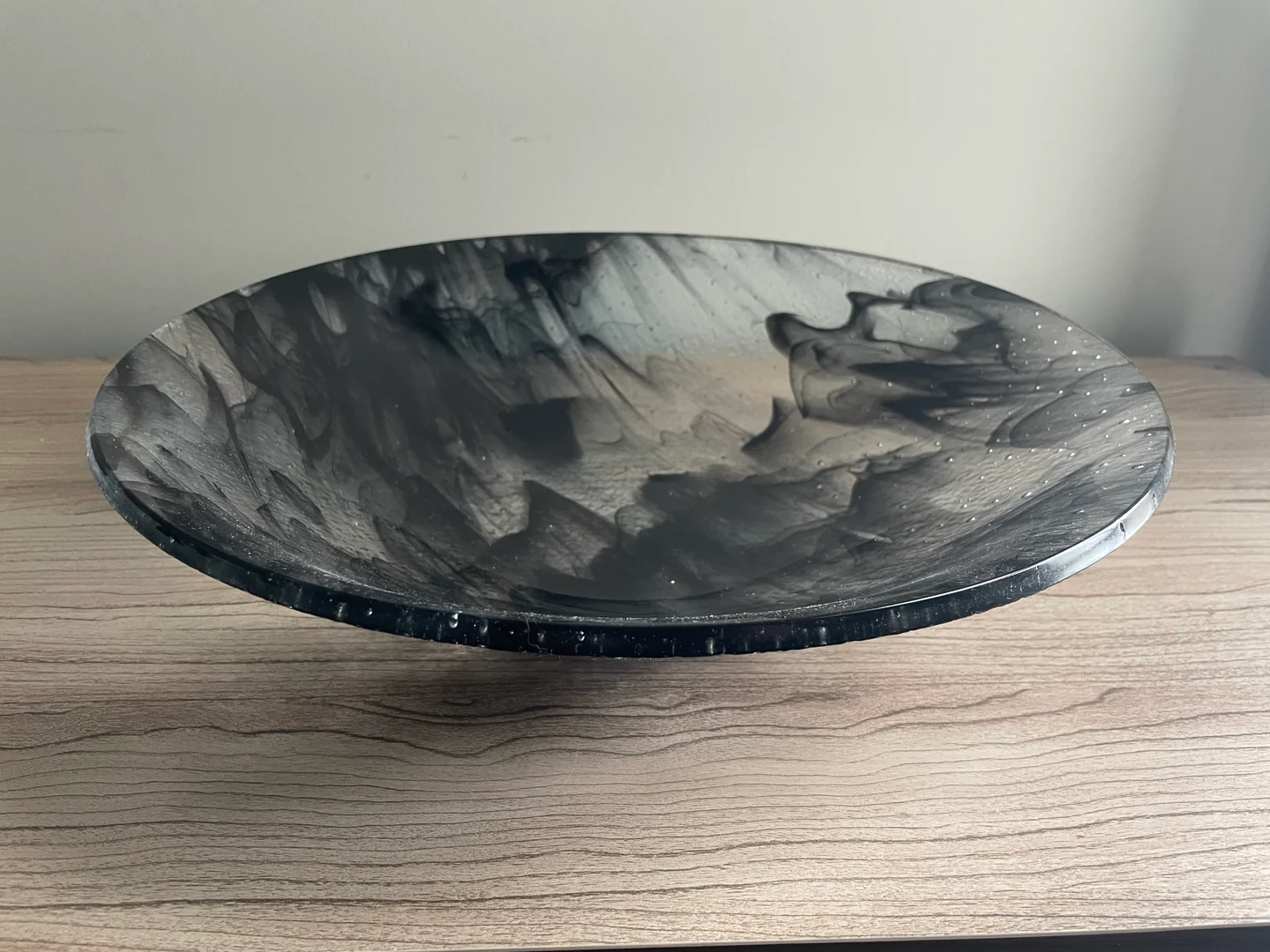 A shallow round bowl with swirling gray and black markings is seen from above at an angle on a pale wood surface.