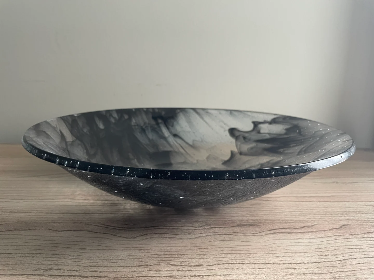 A low side view shows the wide rim and dark underside of a gray-and-black swirled bowl on a pale wood surface.