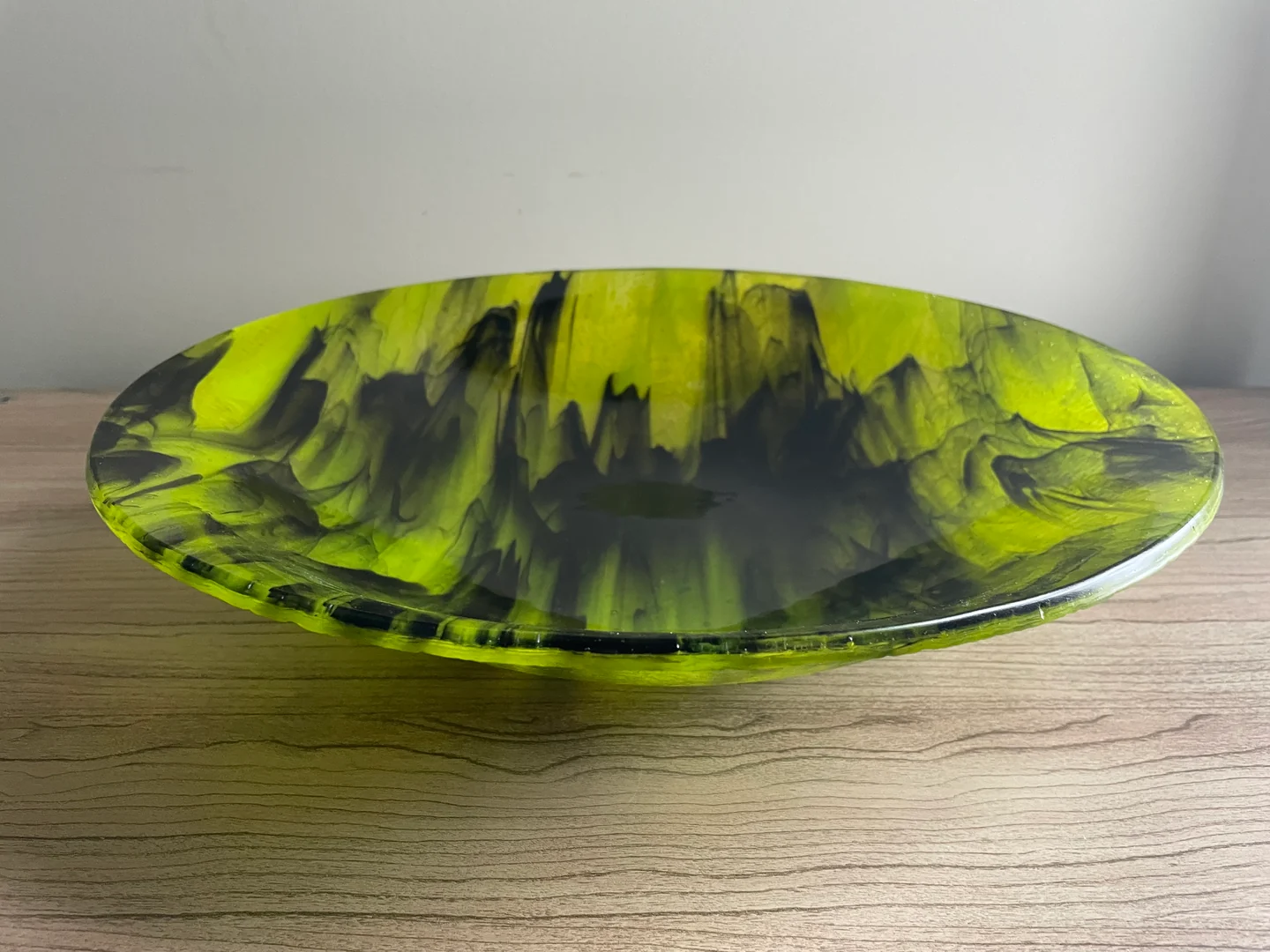 A shallow round bowl with vivid yellow-green and charcoal markings is seen at an angle on a pale wood surface.