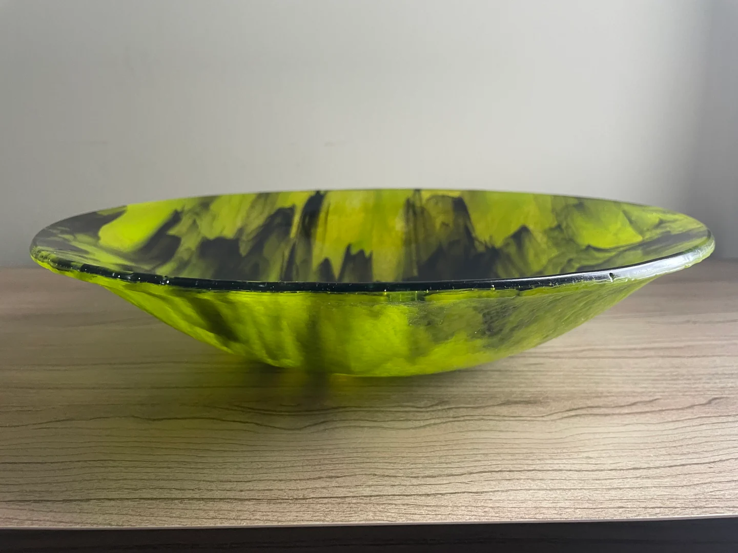 A side view of a low yellow-green and charcoal bowl shows its thin, wide rim above a pale wood surface.
