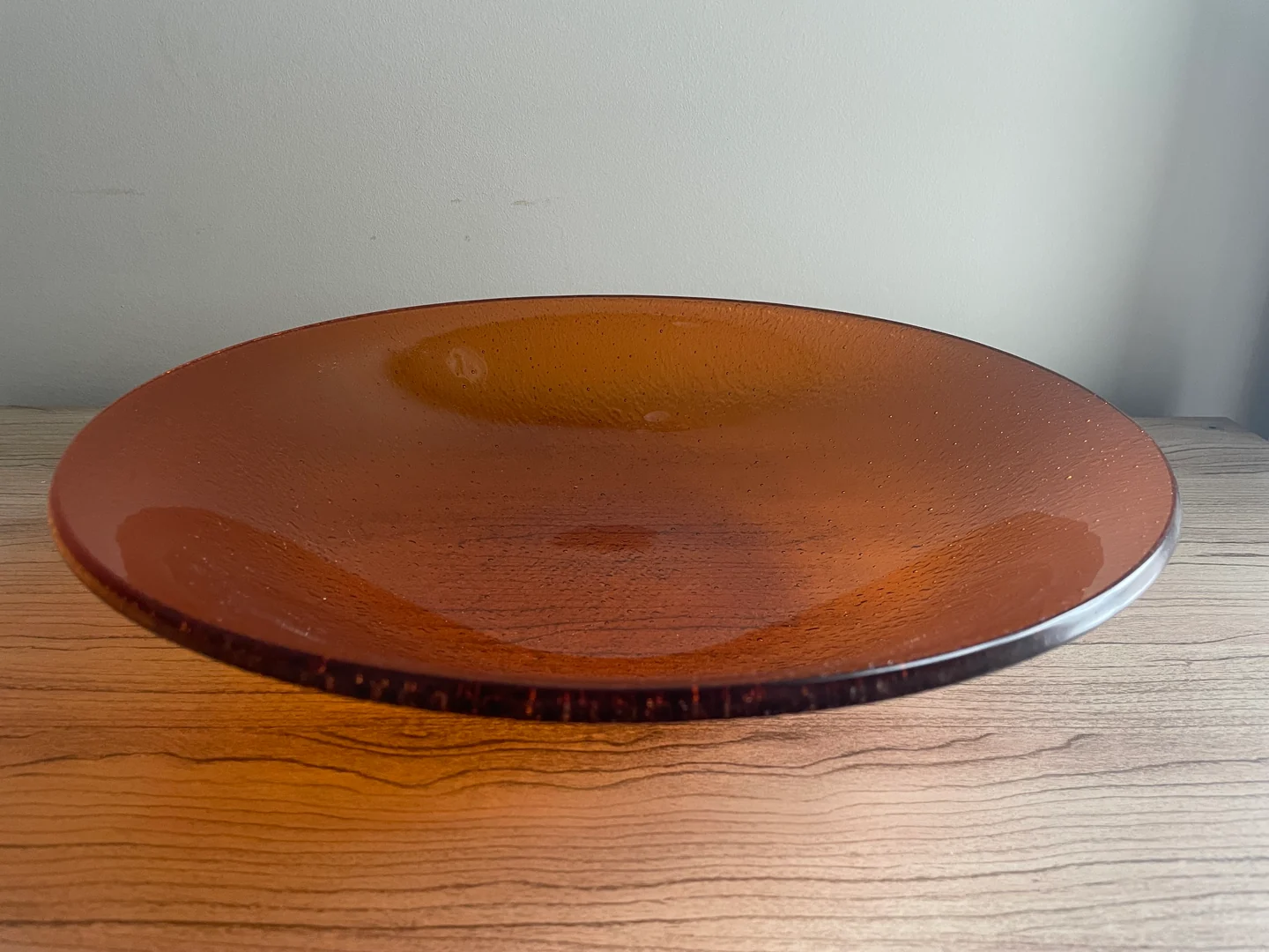 A wide, shallow rust-orange bowl with a lighter amber circle near its center sits on a pale wood surface.