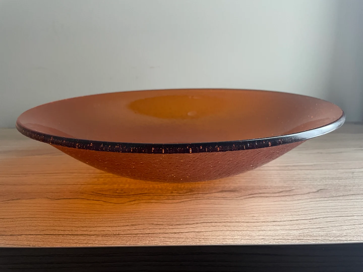 A low side view of a broad rust-orange bowl shows its raised center and dark edge on a pale wood surface.