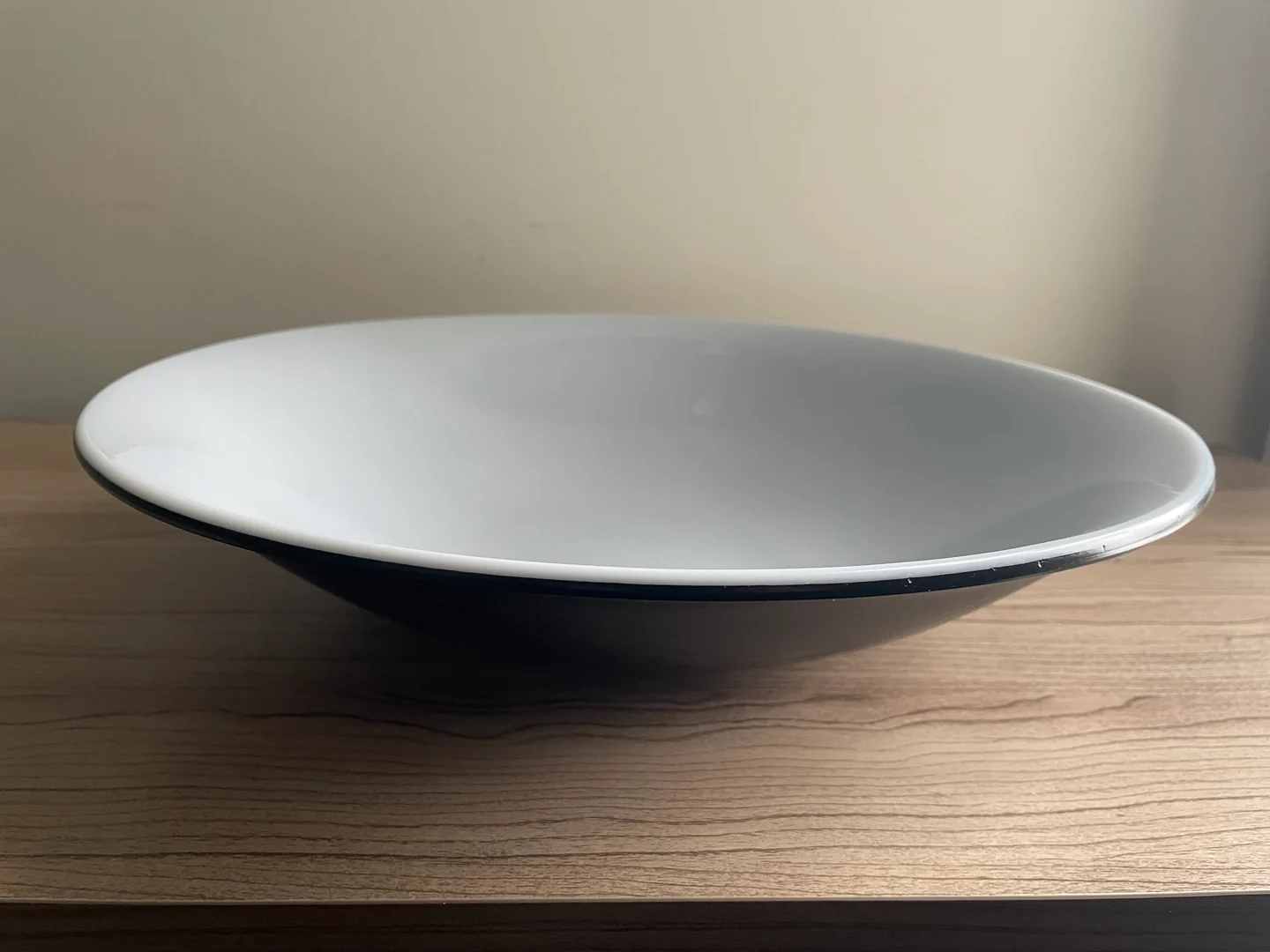 A broad shallow bowl with a pale white interior and dark underside sits on a light wood surface.