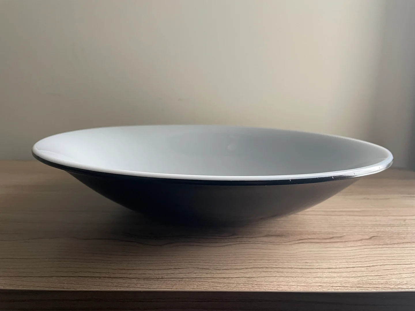 A low side view shows the thin pale rim and dark underside of a wide white bowl on a light wood surface.