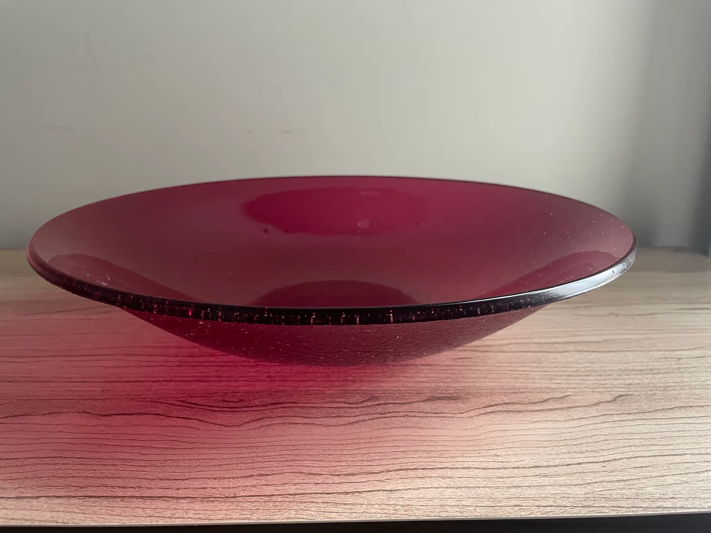 A low side view of a wide wine-red bowl shows its circular hollow and darker rim above a pale wood surface.