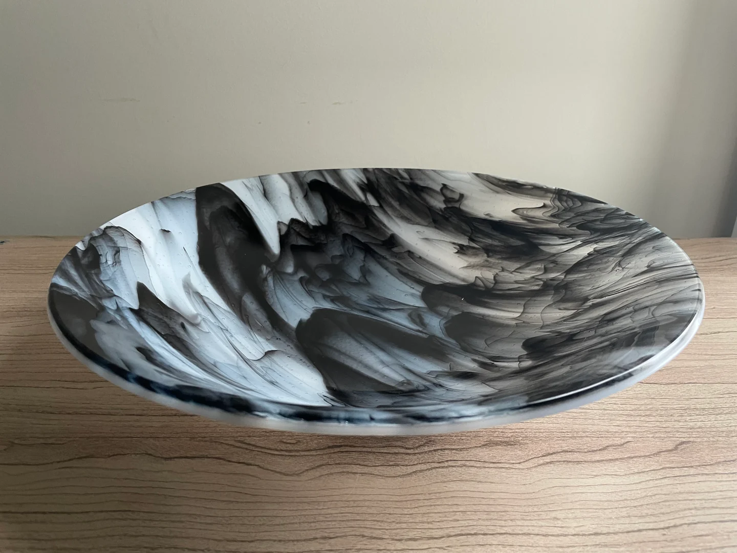 A round shallow bowl shows sweeping black, white and gray bands across its interior on a pale wood surface.