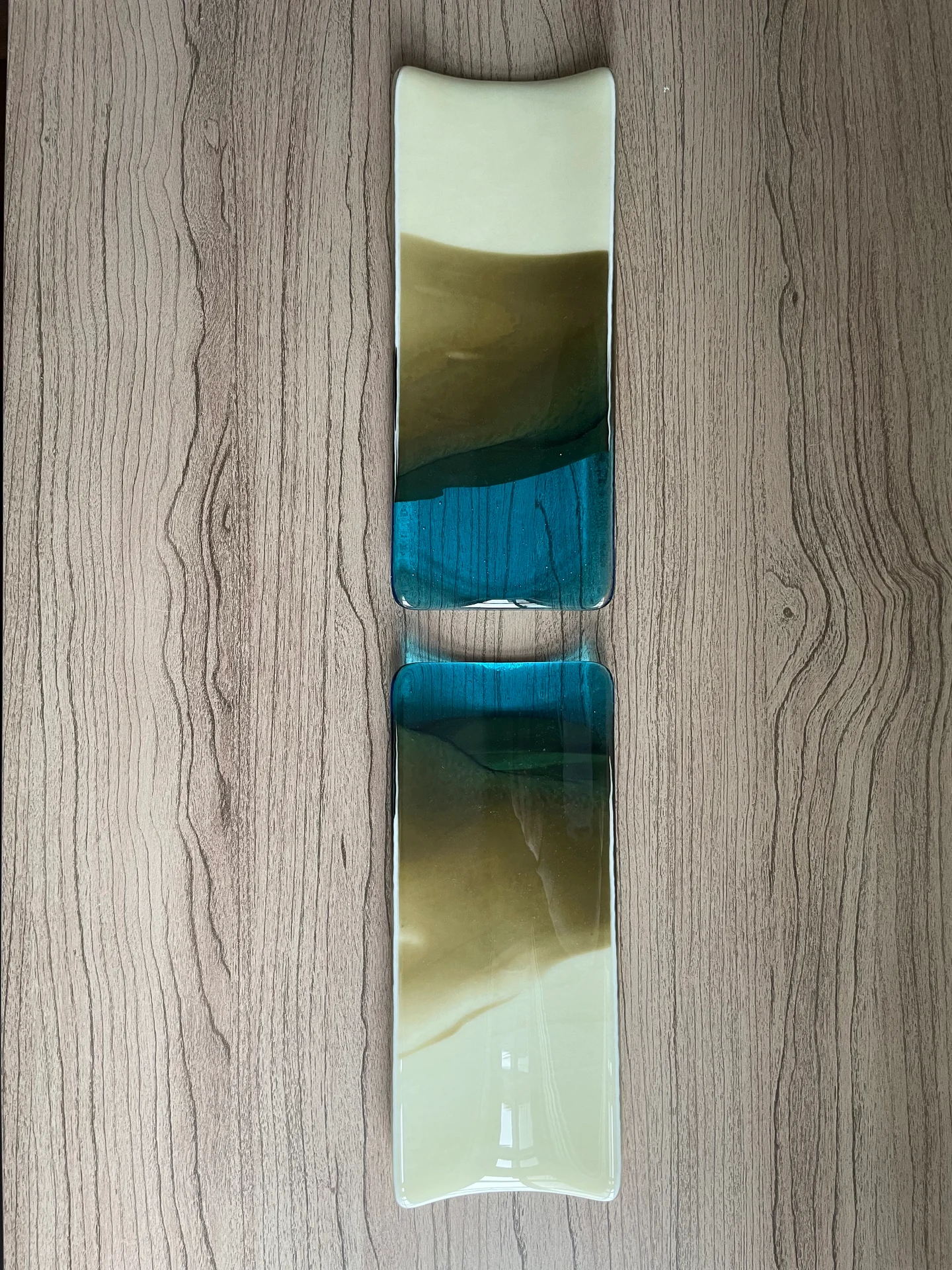 Two separate narrow curved-end forms lie end to end on a wood-grain surface, fading from cream through olive to turquoise blue.