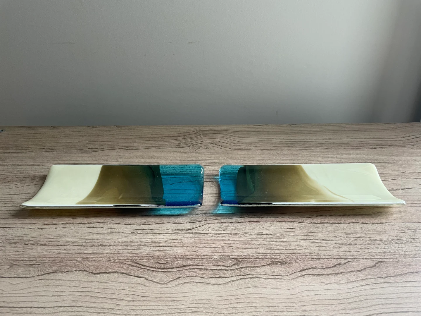 Two long, shallow forms face each other across a narrow gap on a wood-grain surface, each with cream, olive and blue bands.