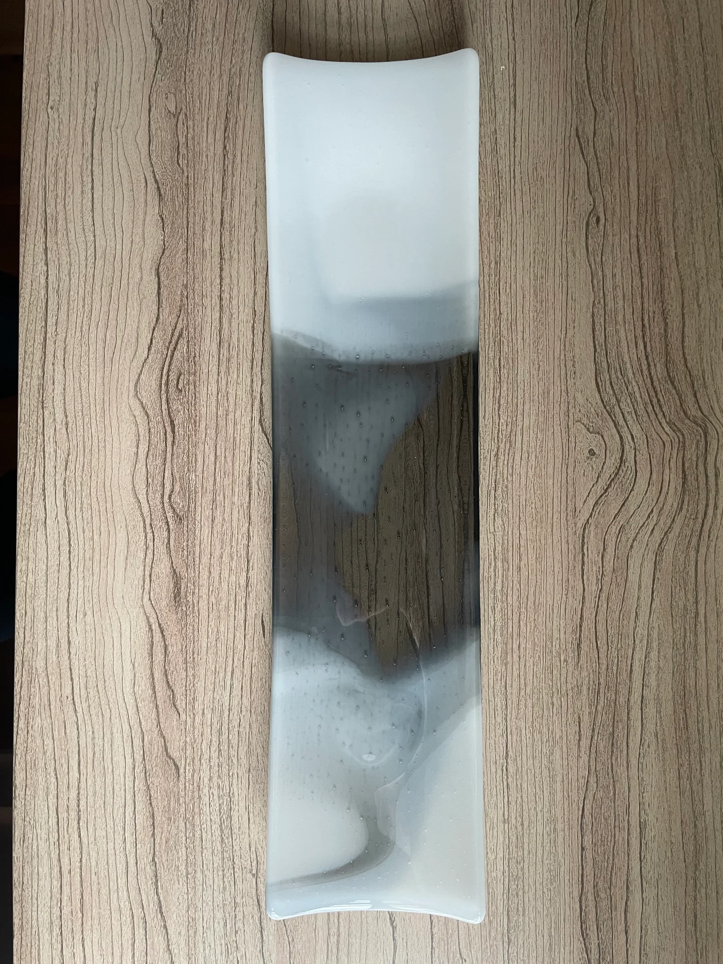 A narrow rectangular form with gently curved ends lies vertically on wood grain, with pale white ends and a smoky dark middle.