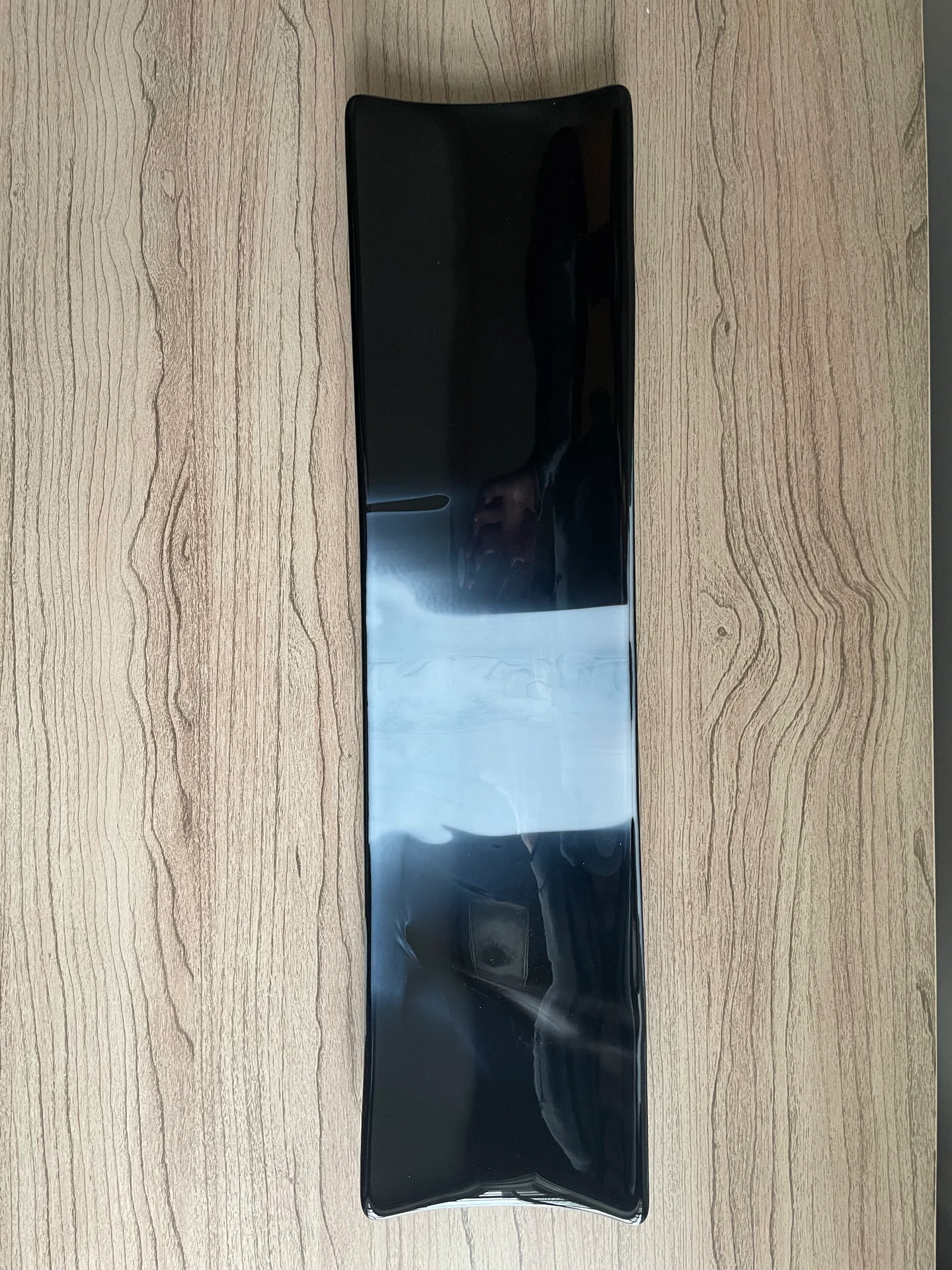 A narrow dark rectangular form with gently curved ends and a wide white band across its center lies vertically on wood grain.