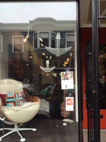 A street-facing window shows white curved grid-like forms above a pale chair, with buildings and a parked car reflected in the glass.